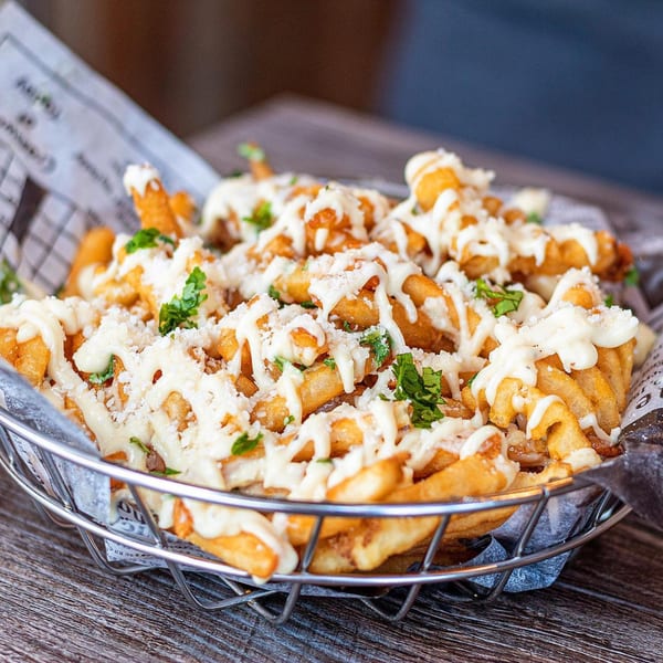 Burbowl's crispy fries topped with creamy white sauce, grated cheese, and fresh parsley in a metal basket.