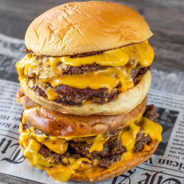 A towering triple-decker Burbowl burger with melted cheese dripping over each patty, on newspaper.