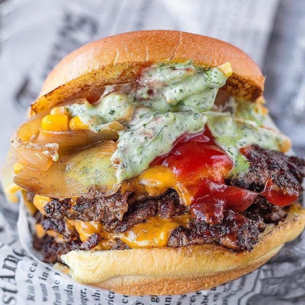 A close-up of a double cheeseburger with grilled onions, corn, ketchup, and green sauce on a bun.