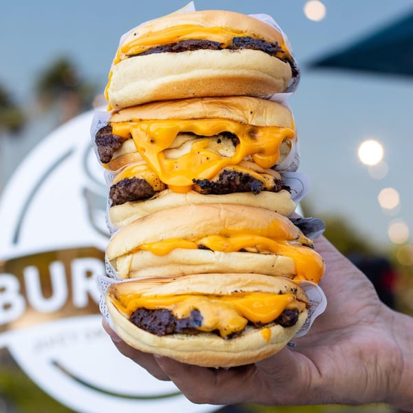 A hand holds a towering stack of five cheeseburgers with melted cheese and beef patties.