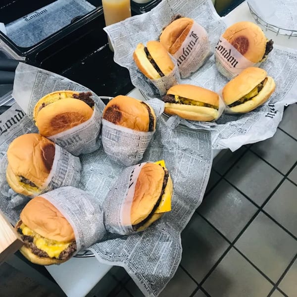 Several cheeseburgers, some wrapped in newspaper-print paper, sit in baskets on a counter.